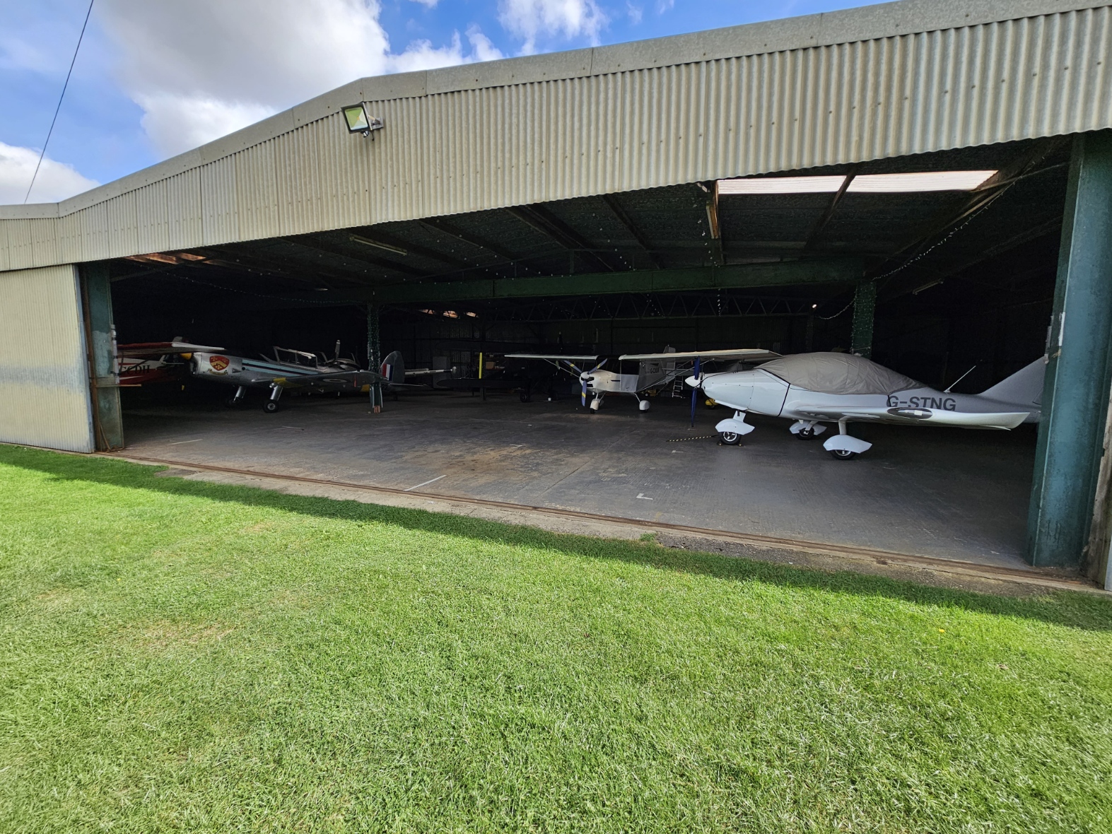 A corrugated iron aircraft hanger. There are five aircrafts visible. On the right a silver painted Chipmunk, with an RAF roundel visible under it's left wing. On the right a white single propeller 3-axis microlight with a cover over the canopy.
